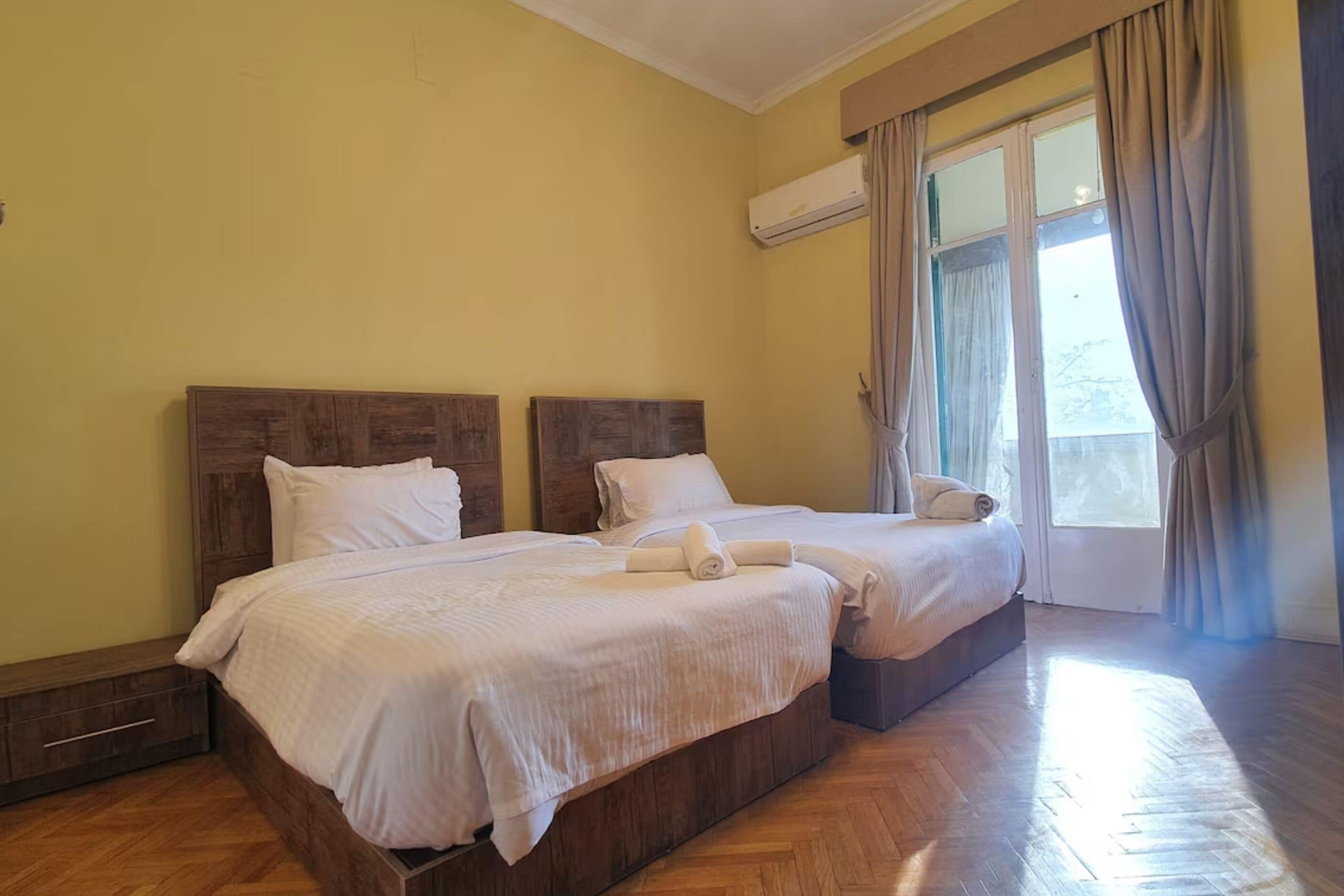 A hotel room with two beds, wooden headboards, a window with curtains, and an air conditioning unit.
