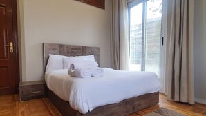 Una cama bien tendida con sábanas y toallas blancas, un cabecero de madera y vista a un edificio a través de un ventanal con cortinas.