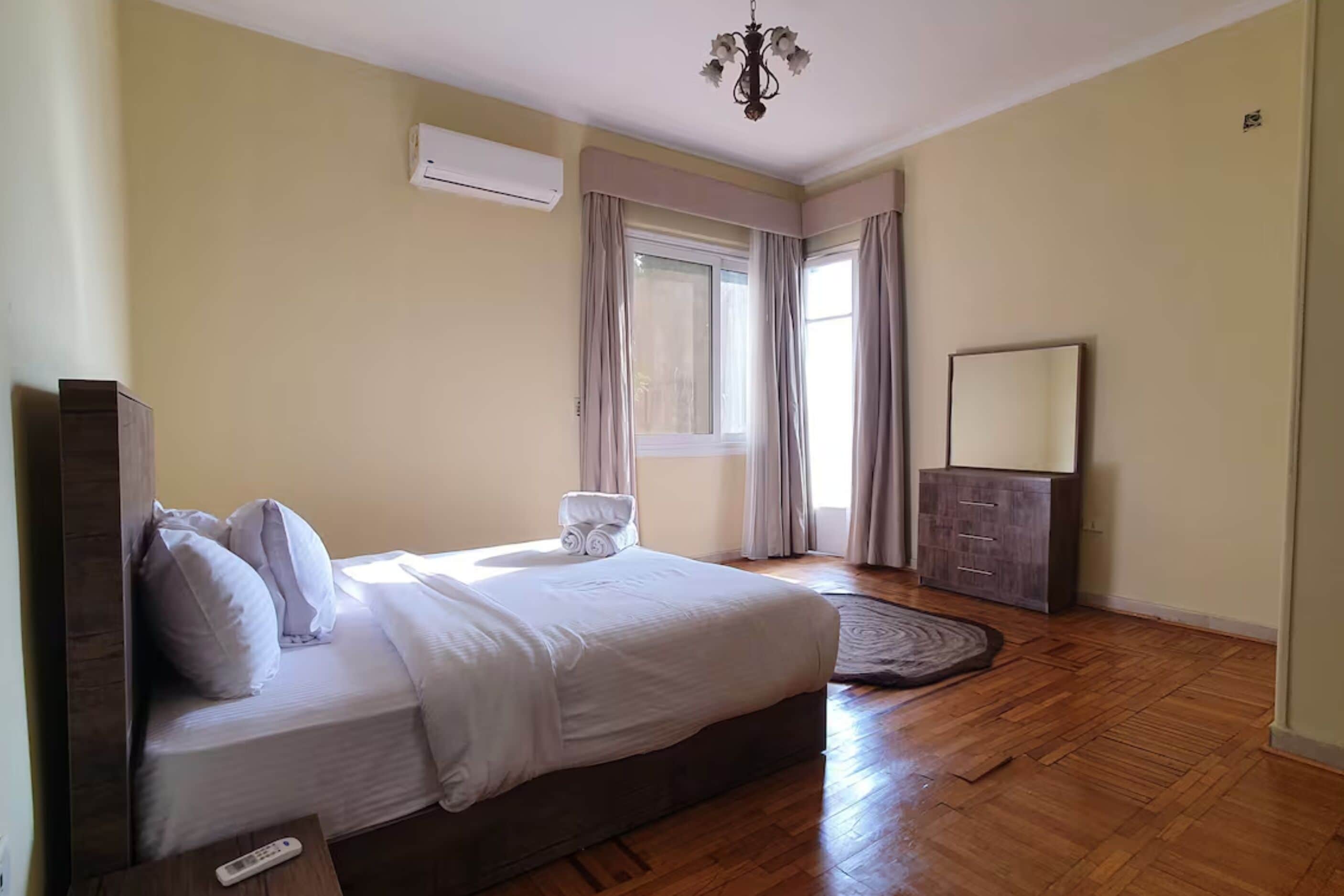 A bedroom with a large bed, a bedside table with a remote control, a wardrobe, and a window with curtains.