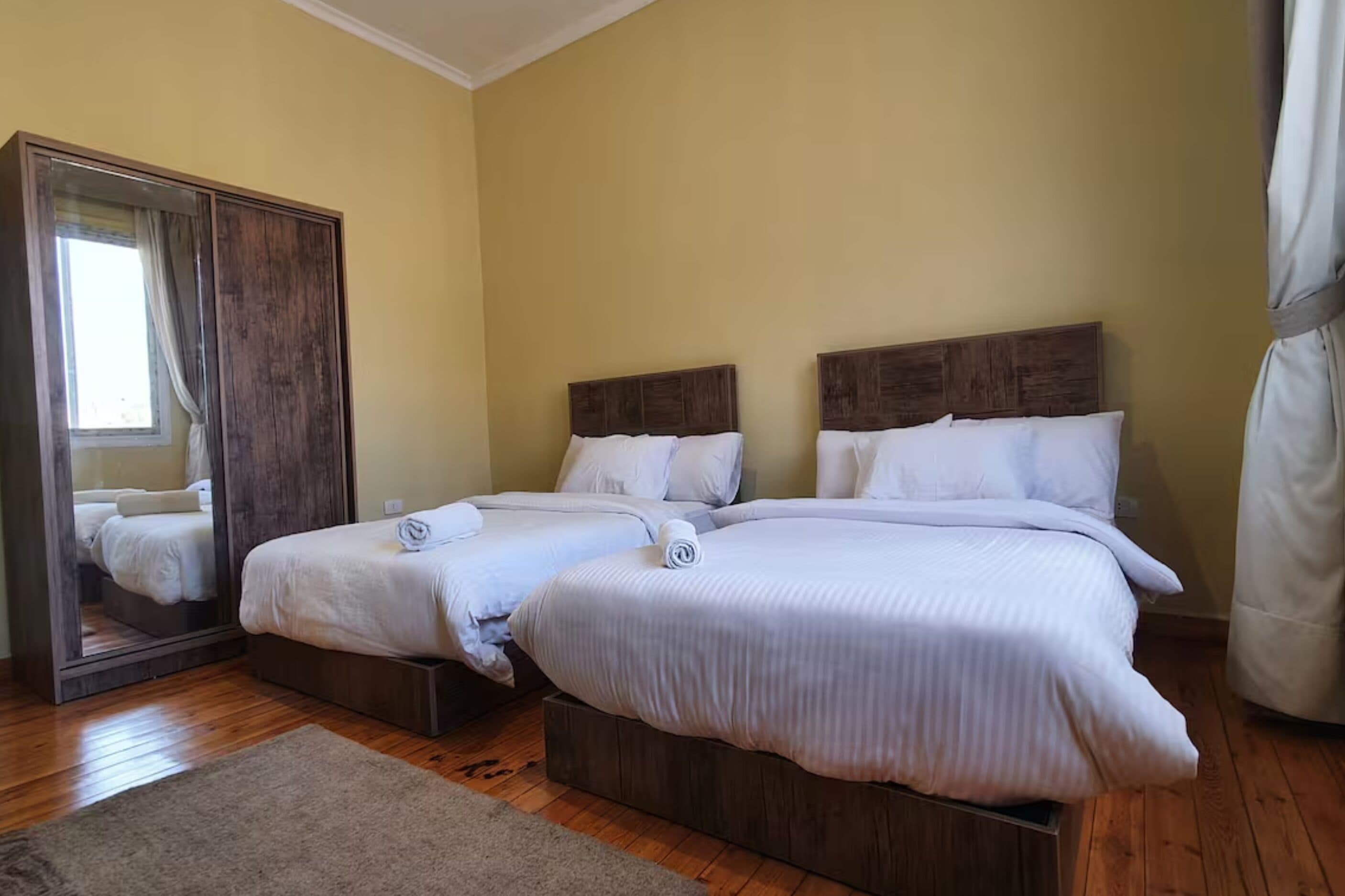 Une chambre d’hôtel avec deux lits, un plancher en bois, un grand miroir et un mur de couleur beige.