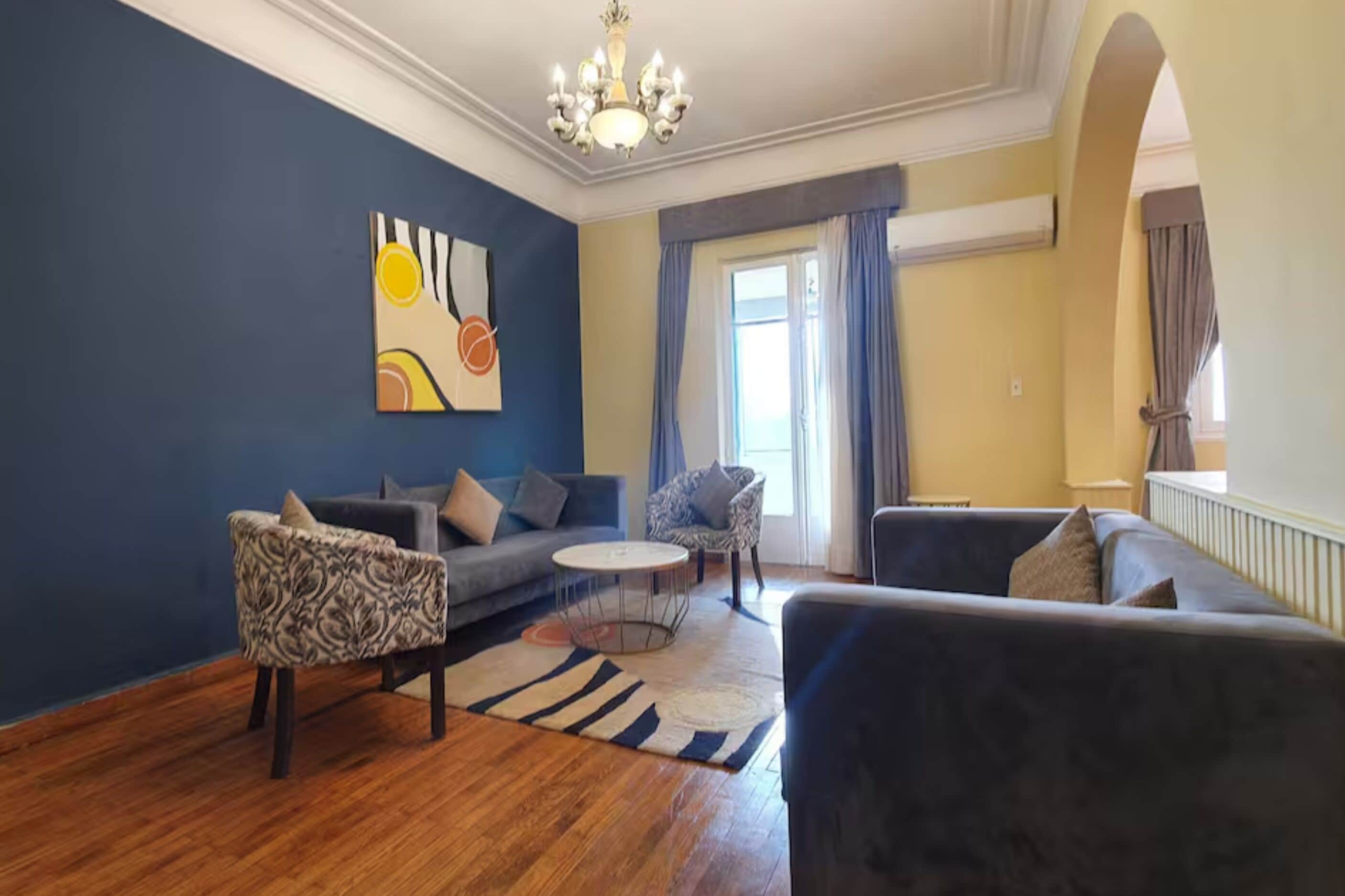 Una sala de estar con pared azul, una araña, un sofá, un sillón estampado y una mesa de centro redonda.