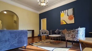 Una sala de estar con un sofá azul, un sillón estampado y una mesa de centro redonda. Se observa una araña, un cuadro en la pared y una alfombra en el suelo.