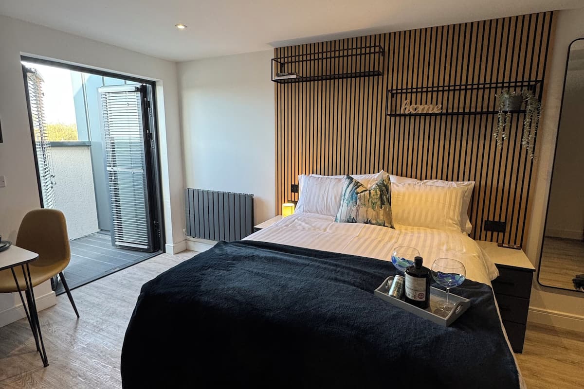 A modern bedroom with a large bed, a bedside table with a bottle and glasses, a chair, and a view of the outdoors through a sliding door.