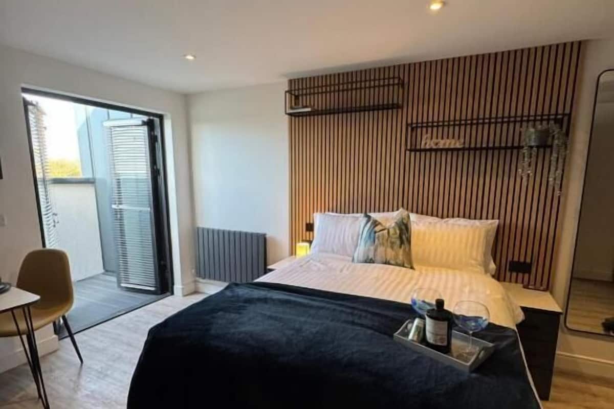 A modern bedroom with a large bed, a desk, and a sliding glass door leading to a balcony.