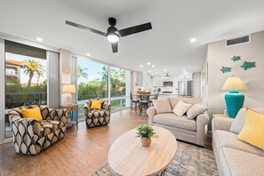 A spacious living area with a ceiling fan, large windows, and a view of palm trees.