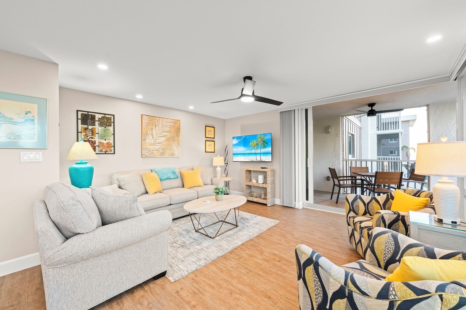 A modern living room with a grey sofa, a coffee table, and patterned armchairs. There is a TV, a ceiling fan, and artwork on the walls.
