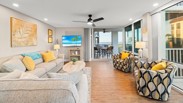 Ein modernes Wohnzimmer mit einer Couch, Sesseln, einem Couchtisch und einem Fernseher. Es gibt einen Balkon mit Blick aufs Meer.