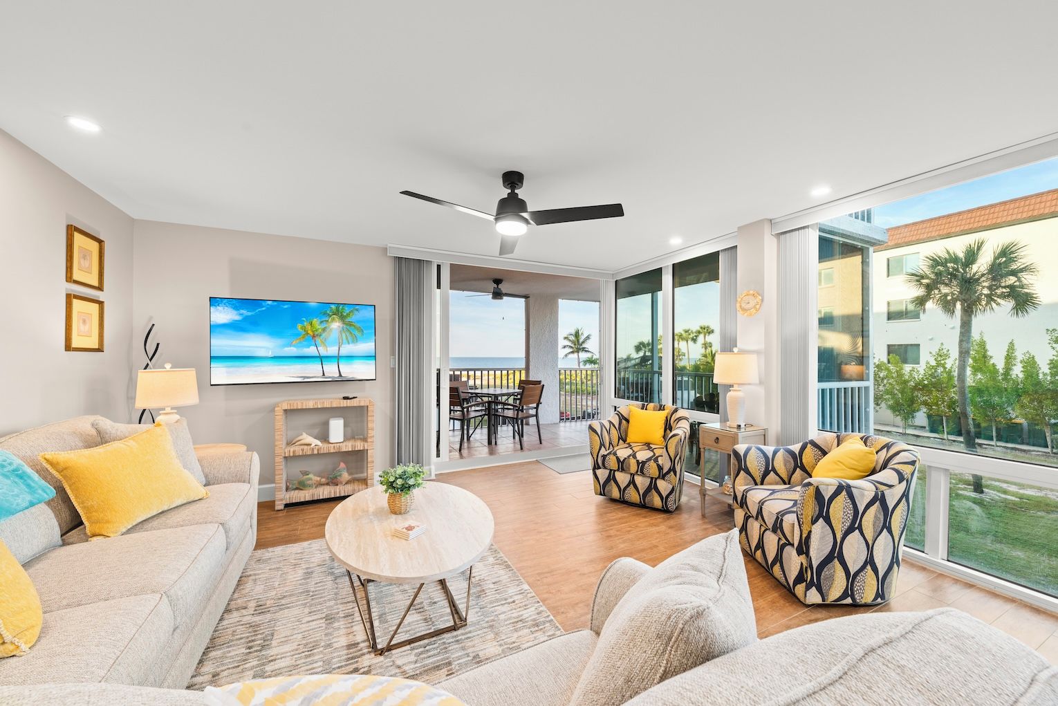 A spacious living room with a large TV, comfortable seating, a balcony with a view of palm trees, and a ceiling fan.