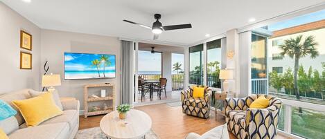 A spacious living room with a large TV, comfortable seating, a balcony with a view of palm trees, and a ceiling fan.