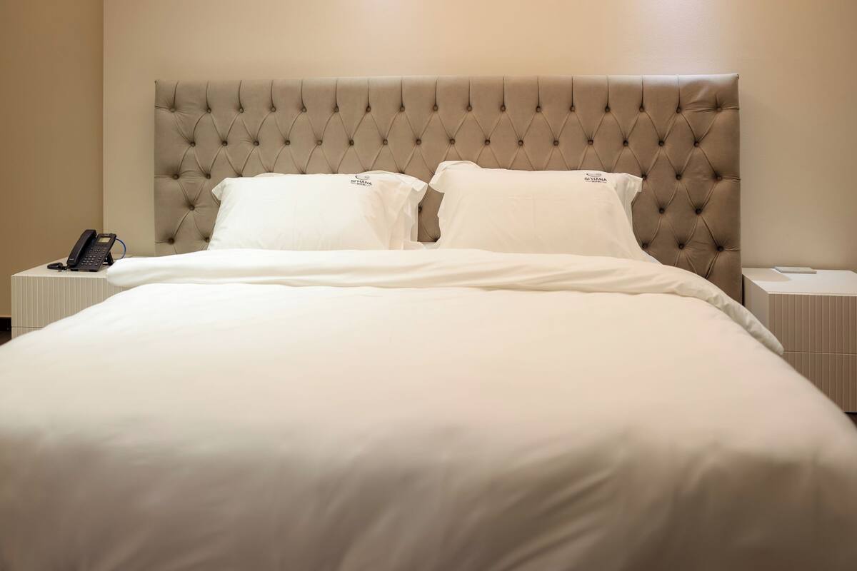 Una cama bien hecha con cabecera capitonada, ropa de cama blanca y un teléfono en la mesita de noche.