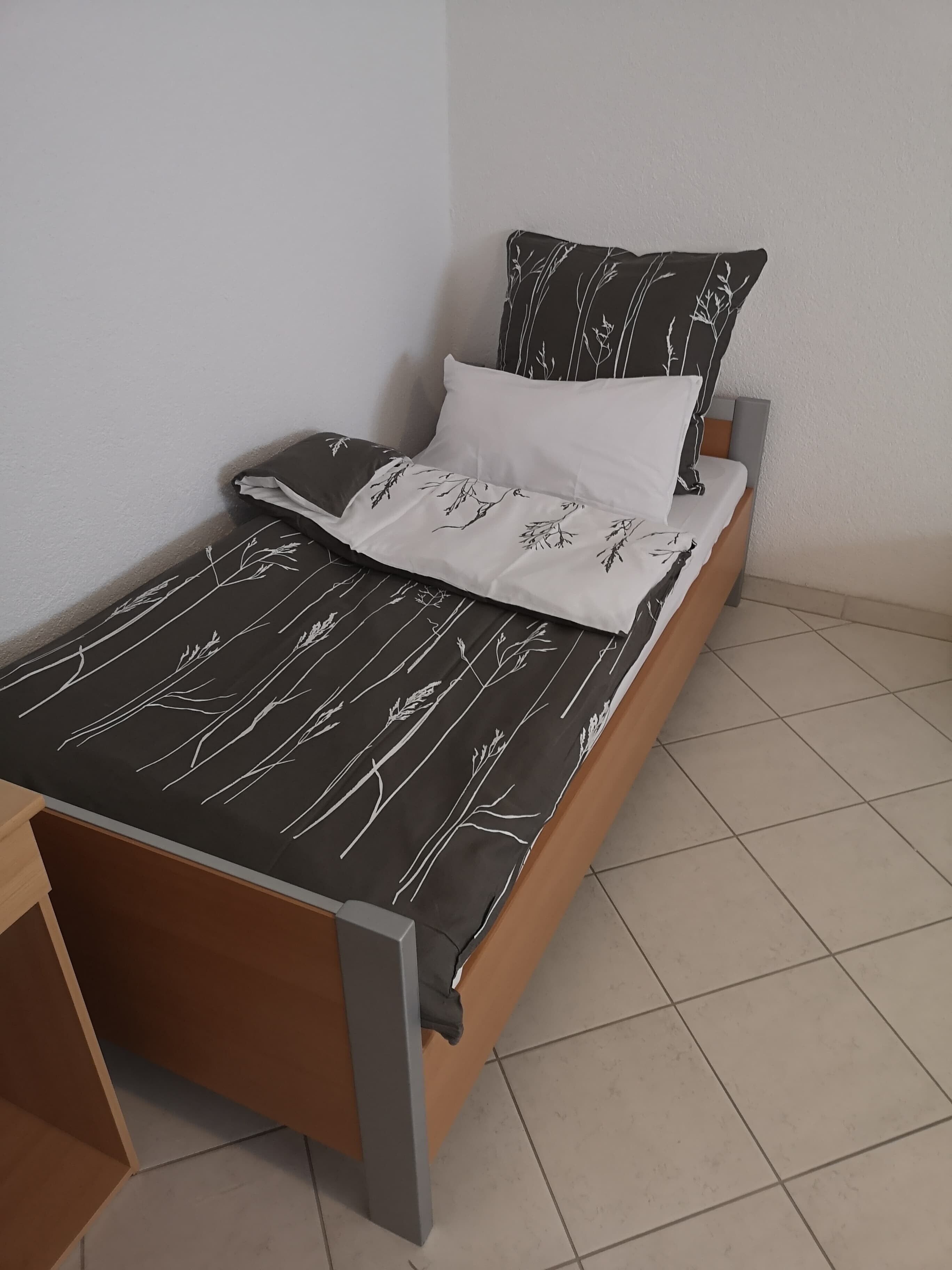 Uma cama de solteiro com colcha estampada em preto e branco, um travesseiro branco e um travesseiro preto com desenho de planta.