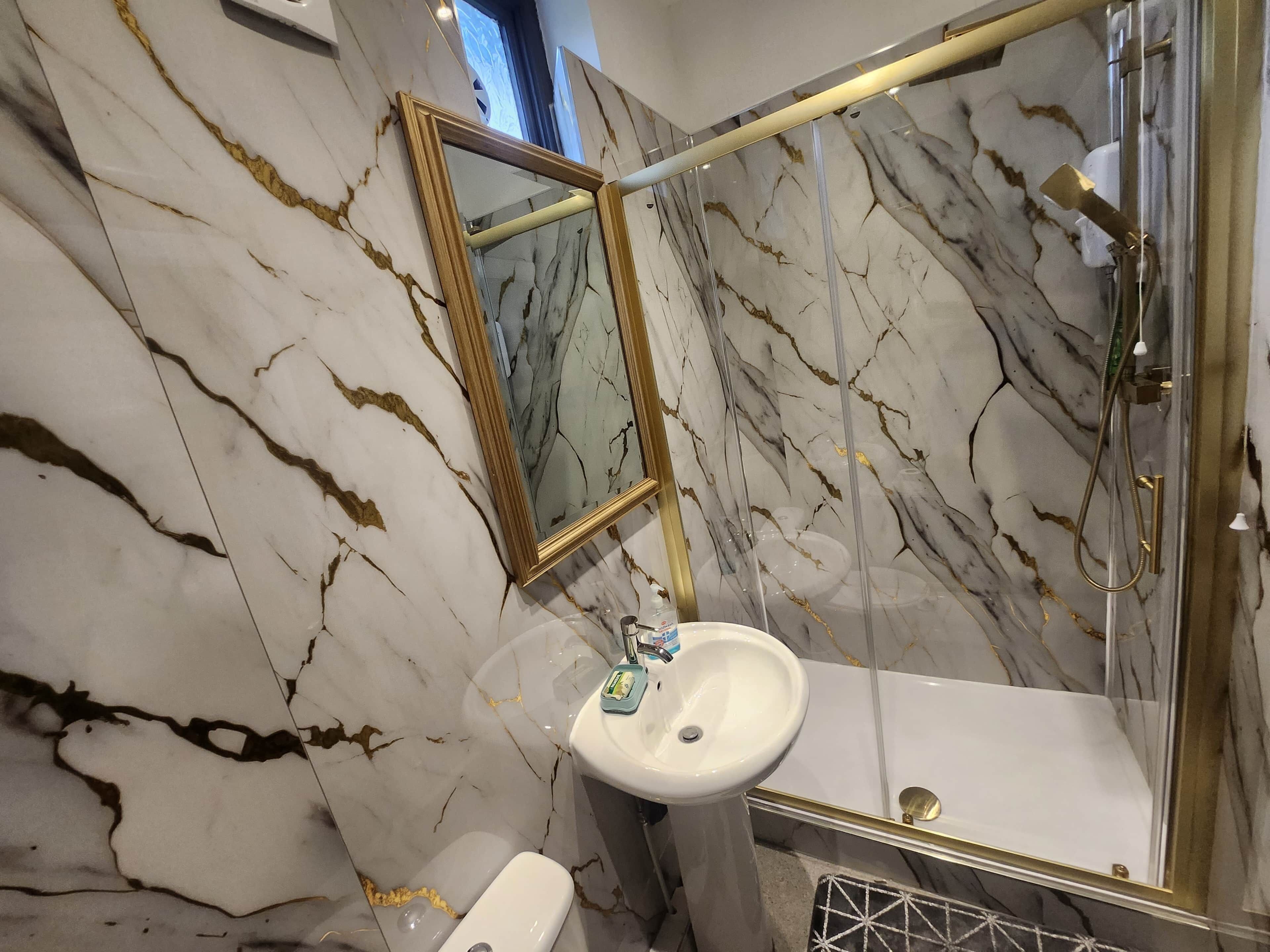 Een badkamer met marmeren wanden, een spiegel met gouden lijst, een wit wastafelmeubel en een douchecabine met glazen wanden.
