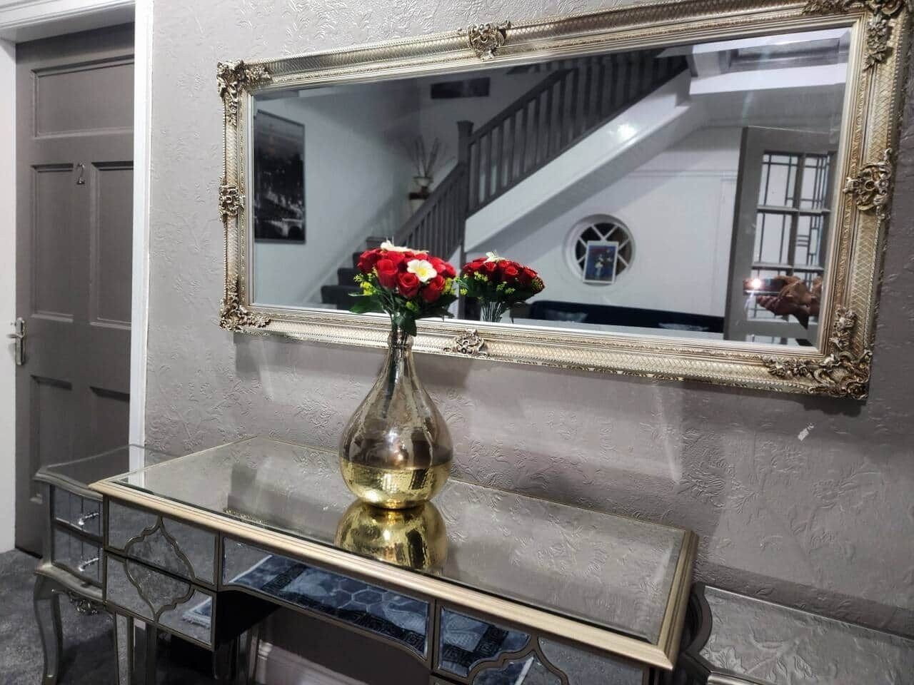 Un miroir avec un cadre doré orné, une console en verre avec un pied doré et un vase rempli de fleurs rouges.