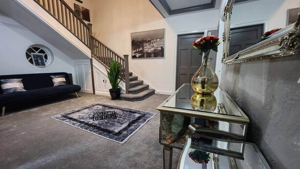 Ein großzügiger Wohnbereich mit Treppe, einem schwarzen Sofa, einem gemusterten Teppich und einem Glastisch mit Blumenvase.