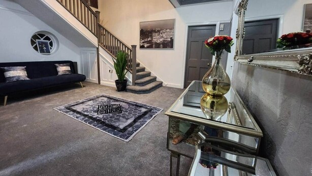 Een ruime woonkamer met een trap, een zwarte bank, een glazen salontafel en een grote spiegel.