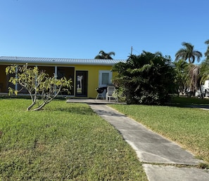 A yellow building with a metal roof, a paved pathway, and a small tree in the foreground. - 3752 San Carlos Drive (Dockside Dreams): 3 BR, 2 BA House in Saint James City, Sleeps 7 (Saint James City)