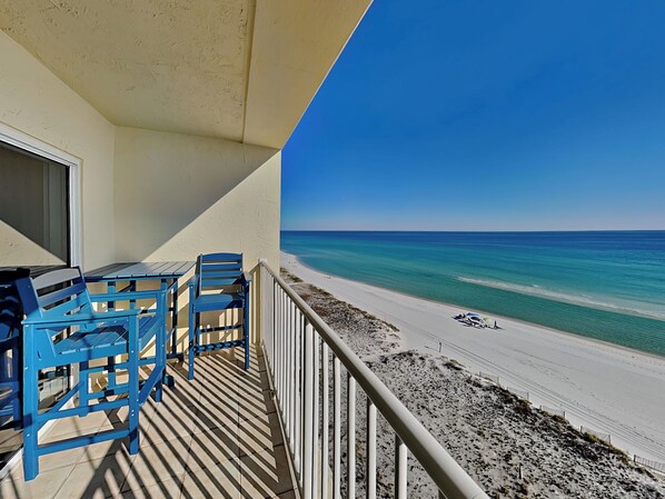 A balcony with blue chairs and a table overlooking a beach with clear blue water and a sandy shore.