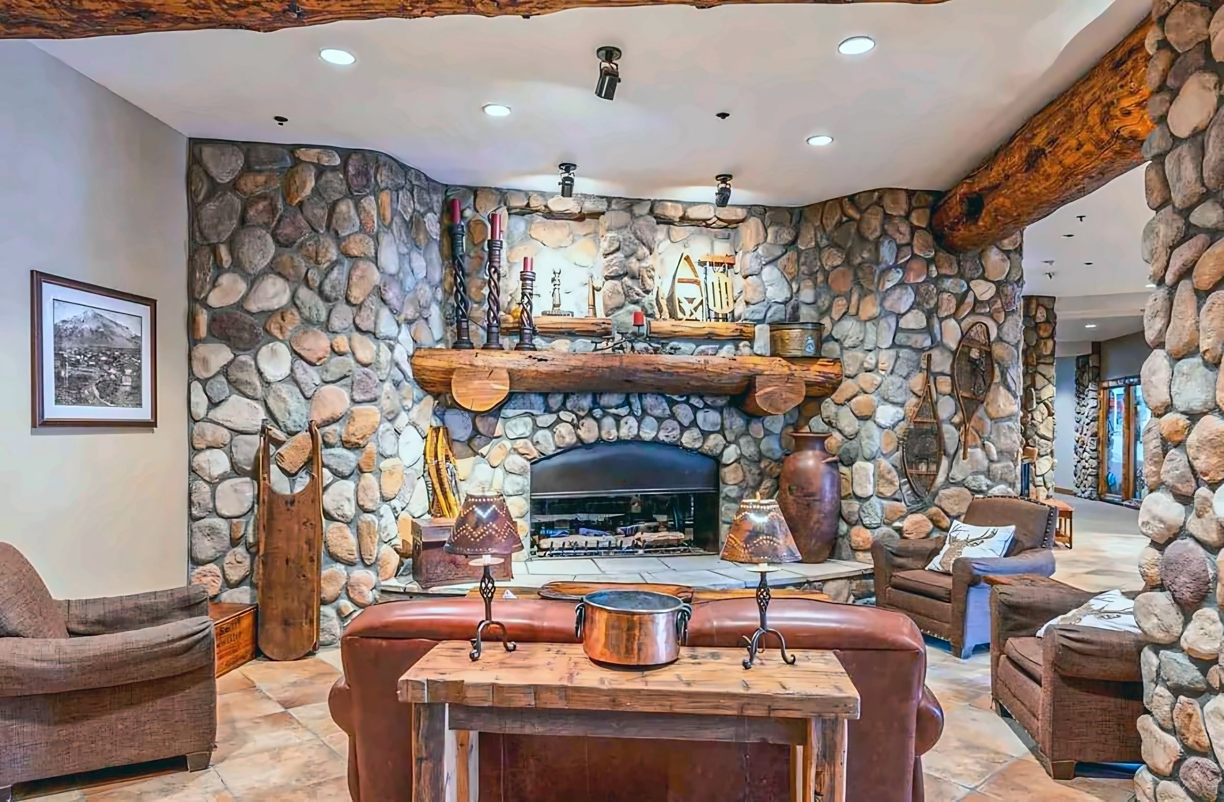 Une cheminée en pierre avec un manteau en bois, entourée de fauteuils en cuir et d’un mur en pierre.