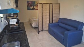 A compact living space with a kitchenette, a blue sofa, a bed, and a decorative tree painting. - Edificio Navarra (Asunción)