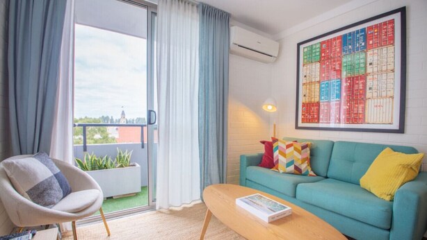 Een moderne woonkamer met een blauwgroene bank, een houten salontafel en een stoel met kussen. Er is een groot raam met gordijnen, een lijstwerk kunstwerk aan de muur en een potplant op het balkon.