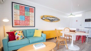 Een moderne woonkamer met een blauwe bank, een houten salontafel en een eethoek met een witte tafel en rode stoelen.