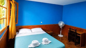 Une chambre avec un lit, un ventilateur, une table de chevet en bois et une fenêtre avec des rideaux.