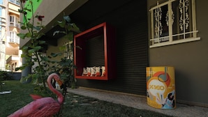 A pink flamingo statue in a garden area with a red shelf displaying bags and a yellow advertisement. - ROTA GEZGİN PANSİYON (Eskisehir)