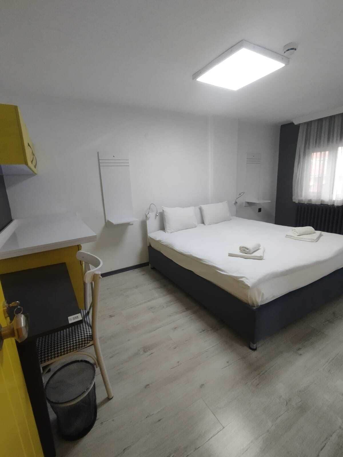 A hotel room with a bed, a desk, a chair, a trash bin, and a window with curtains.