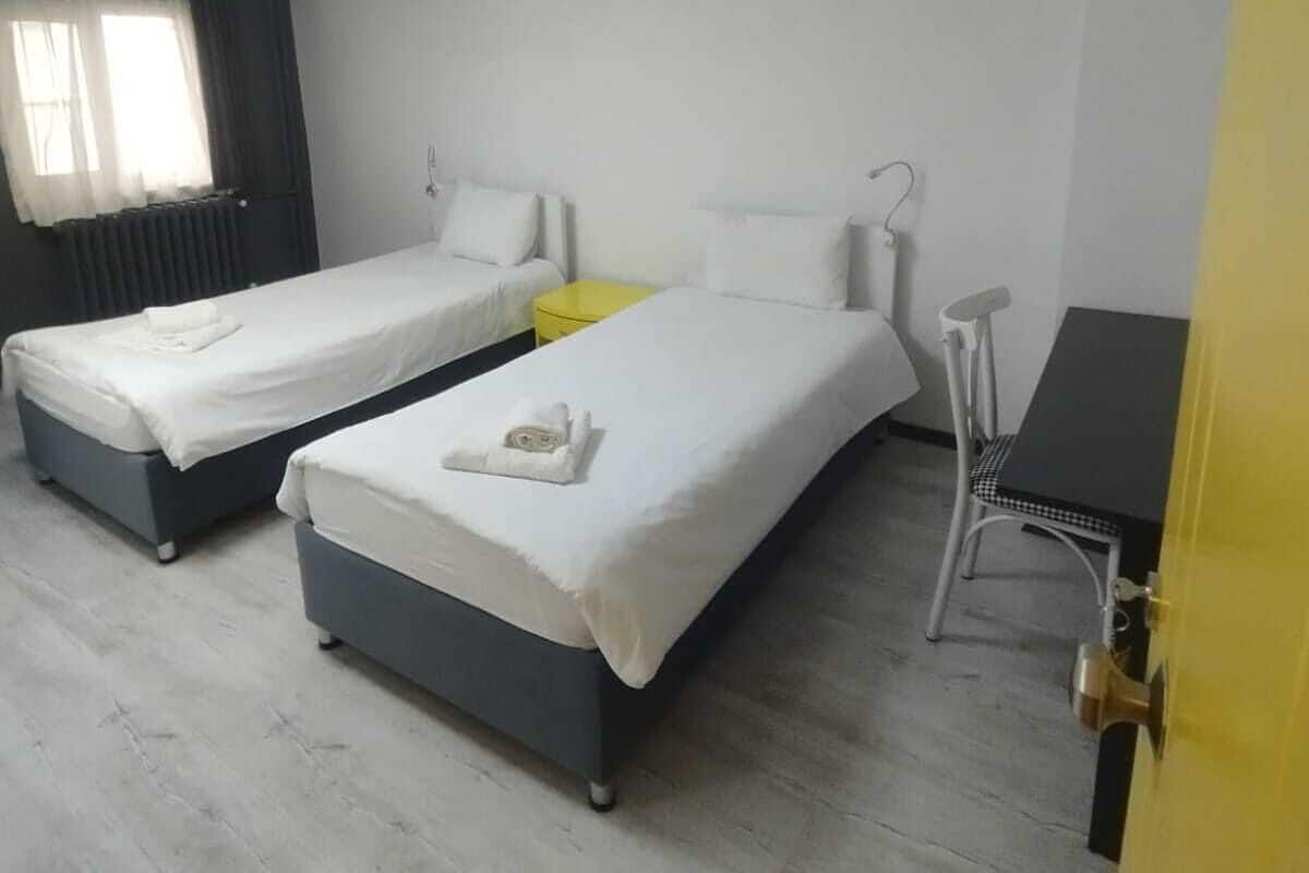 A hotel room with two single beds, a small table, a chair, and a window with curtains.