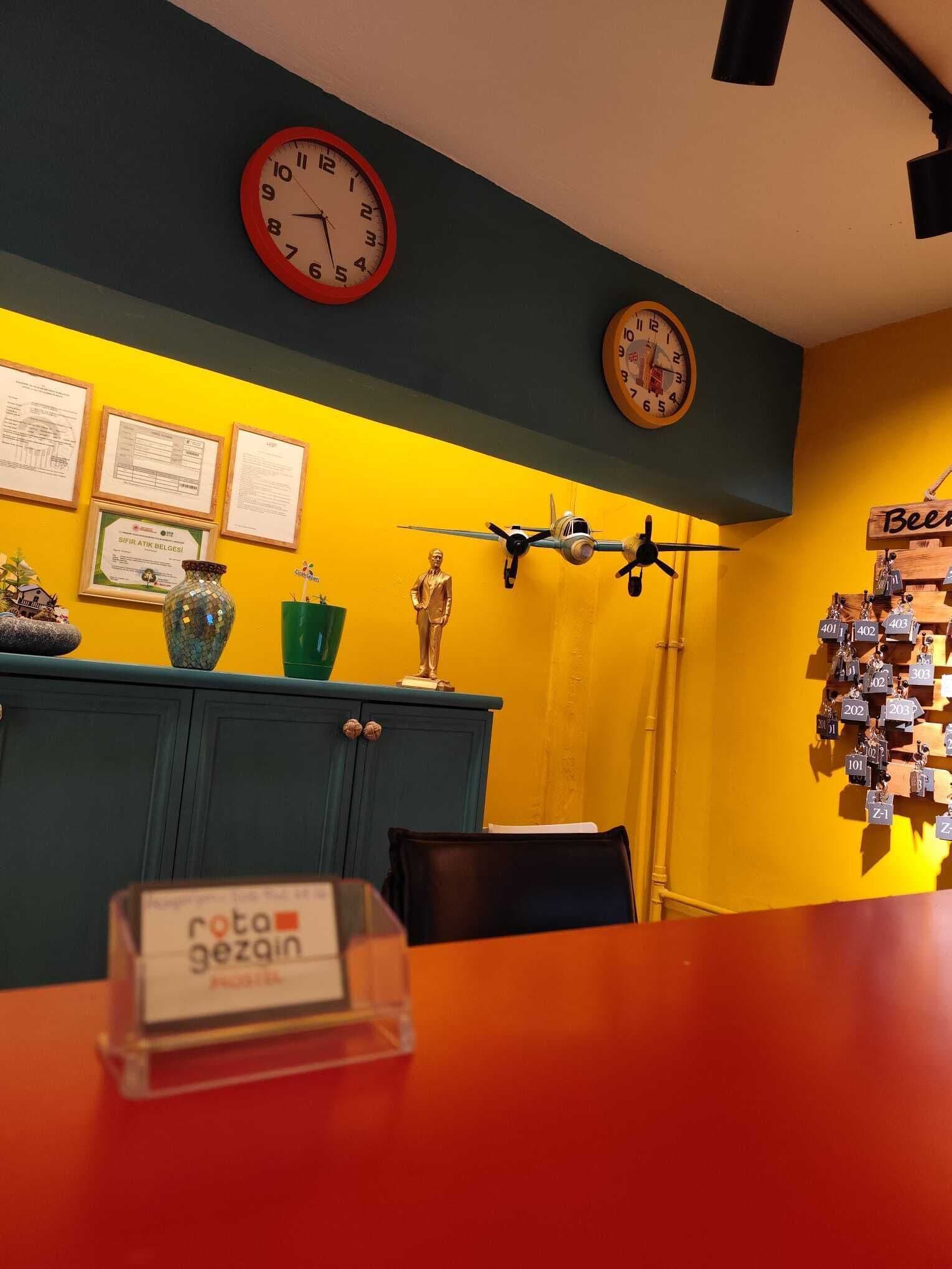 A room with a yellow wall, two clocks, a small airplane model, and a sign reading "rota gezgin".