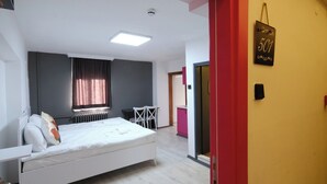A bedroom with a bed, a desk, and a chair. There is a window with curtains, a radiator, and a door with a sign. - ROTA GEZGİN PANSİYON (Eskisehir)