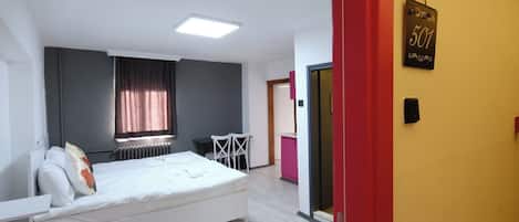 Ein Schlafzimmer mit einem Bett, einem Schreibtisch und einem Stuhl. Es gibt ein Fenster mit Vorhängen, eine Heizung und eine Tür mit einem Schild.