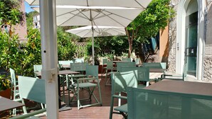 Un patio al aire libre con sillas y mesas de metal verde, cubierto por sombrillas blancas y rodeado de follaje.