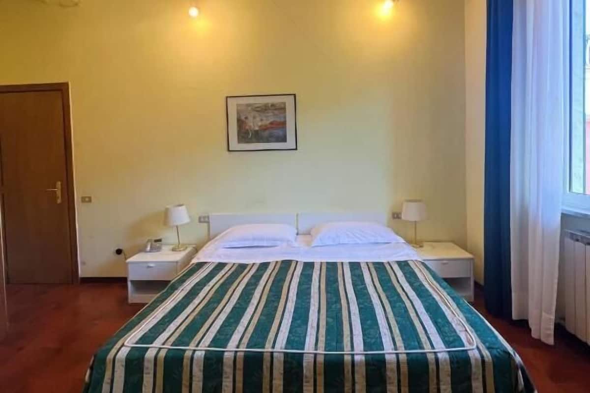 A hotel room with a bed, two bedside tables, a nightstand, a door, and a window with curtains.