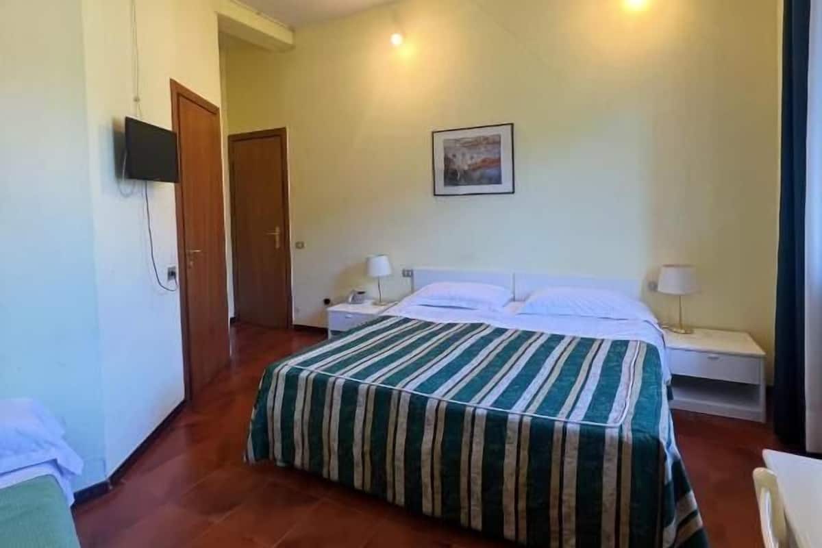 A hotel room with a bed, a bedside table with a lamp, a wall-mounted TV, and a door leading to another room.