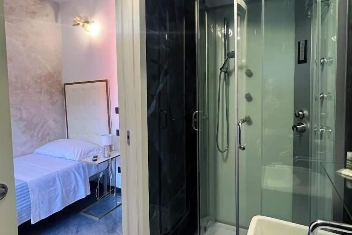 A modern bathroom with a glass-enclosed shower, a white sink, and a bed in the adjacent room.