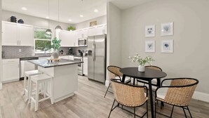 A modern kitchen with white cabinets, stainless steel appliances, a central island, and a dining area with wicker chairs. - Canopy Couplet | 2 Upscale Apts In Prime Location (Safety Harbor)