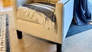 A beige armchair with a striped throw blanket and a pillow, positioned in a well-lit room with a large window and curtains. - 2120  Hot tub, sauna & more resort amenities Beautifully remodeled, 1st-floor (Beech Mountain)