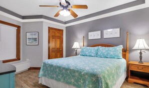 Un dormitorio con una cama, dos mesitas de noche con lámparas, un ventilador de techo, una puerta de madera y cuadros enmarcados en la pared.