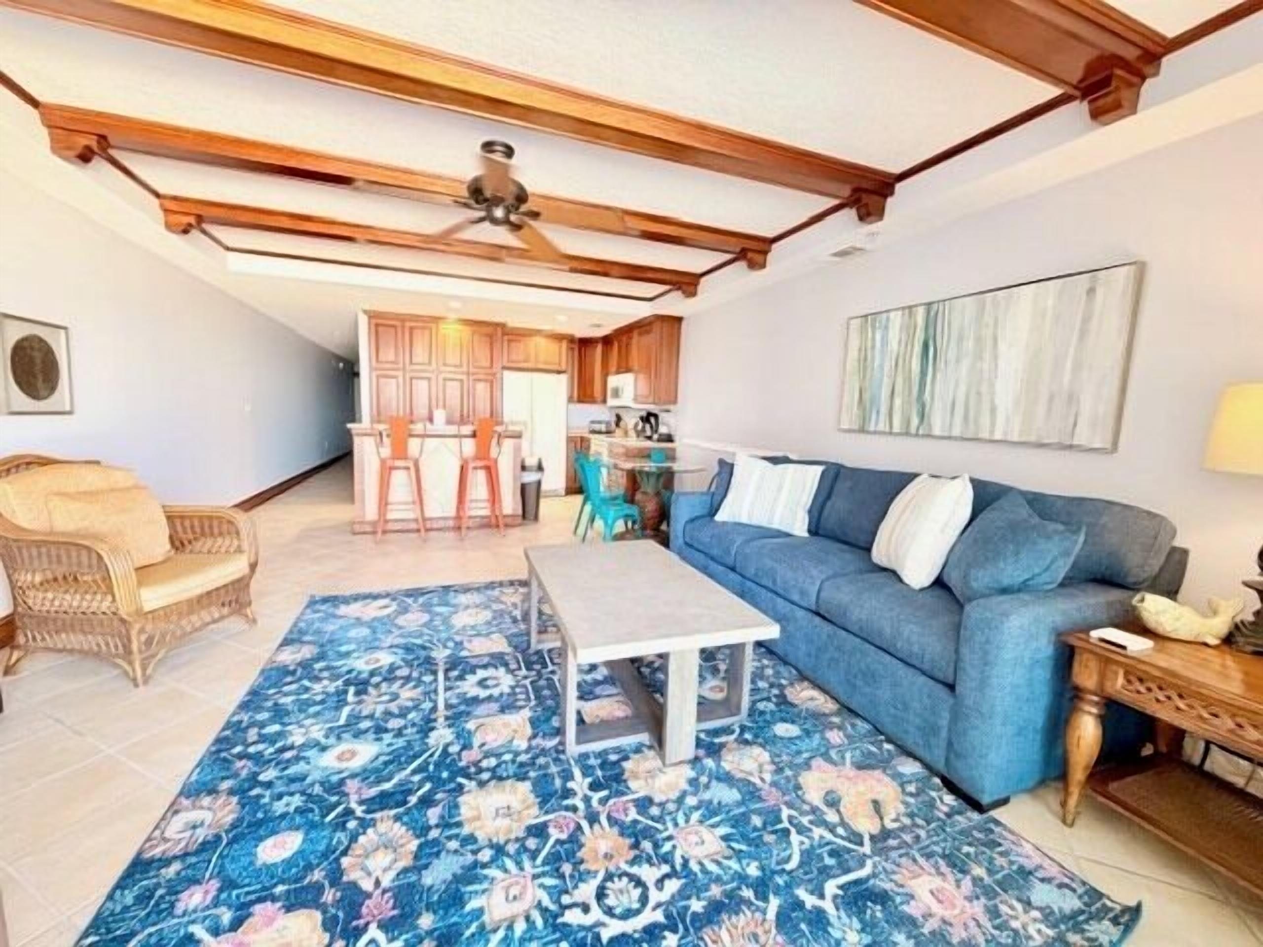 A living room with a blue sofa, a coffee table, and a patterned rug. There is a wicker chair and a wooden table with a lamp. The room has wooden beams on the ceiling and a ceiling fan.
