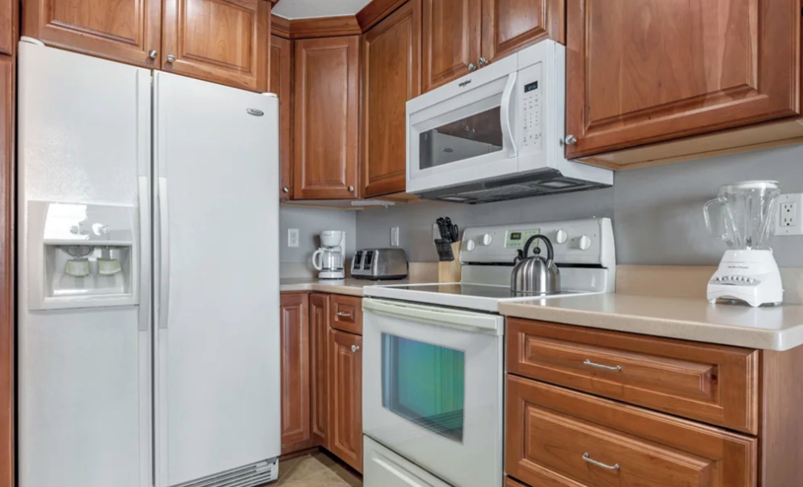 A kitchen with wooden cabinets, a white refrigerator, a microwave, a toaster, a kettle, and a blender.