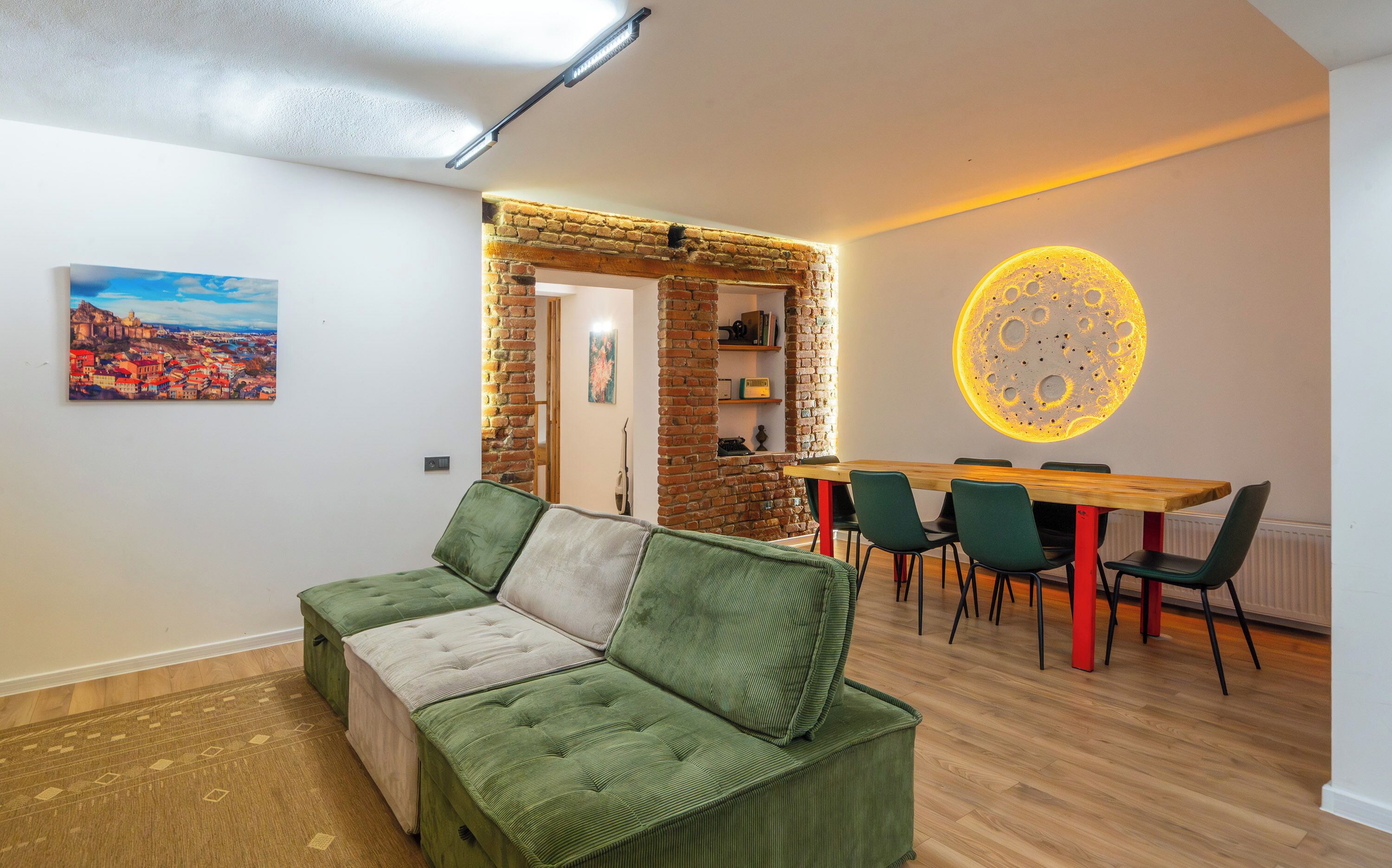 A cozy living room with a green sofa, a dining area with a wooden table and chairs, and a wall with a circular light fixture.