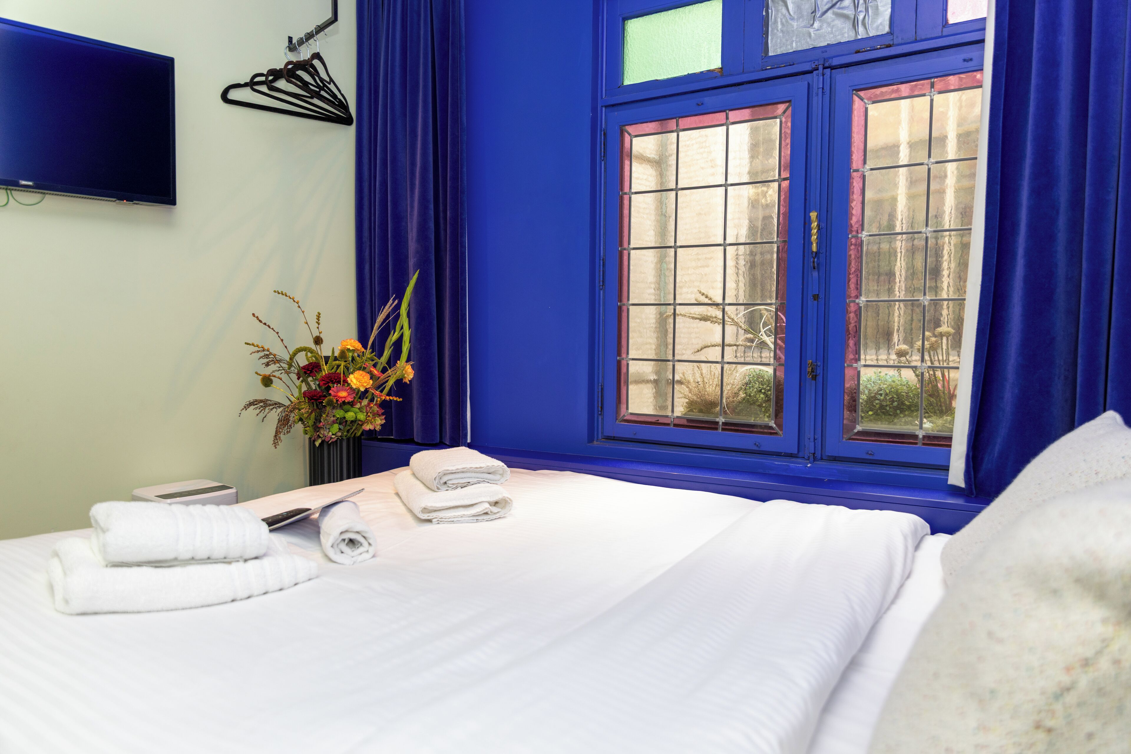 Una habitación de hotel con una cama, un televisor de pantalla plana montado en la pared, una ventana con cortinas azules y un florero con flores en la mesita de noche.