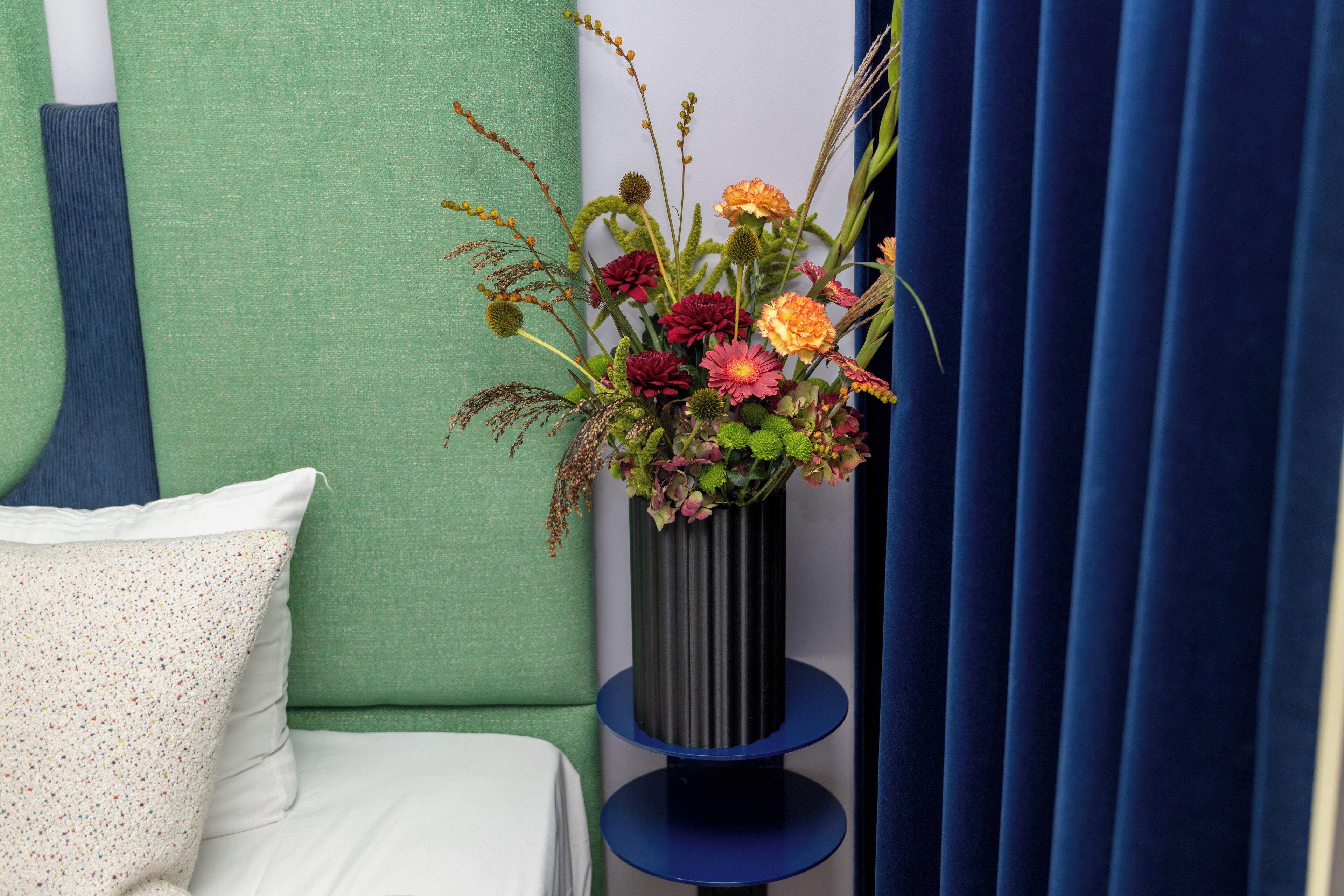 Una cama con ropa de cama blanca y una almohada decorativa, un jarrón alto con flores y una cortina azul.