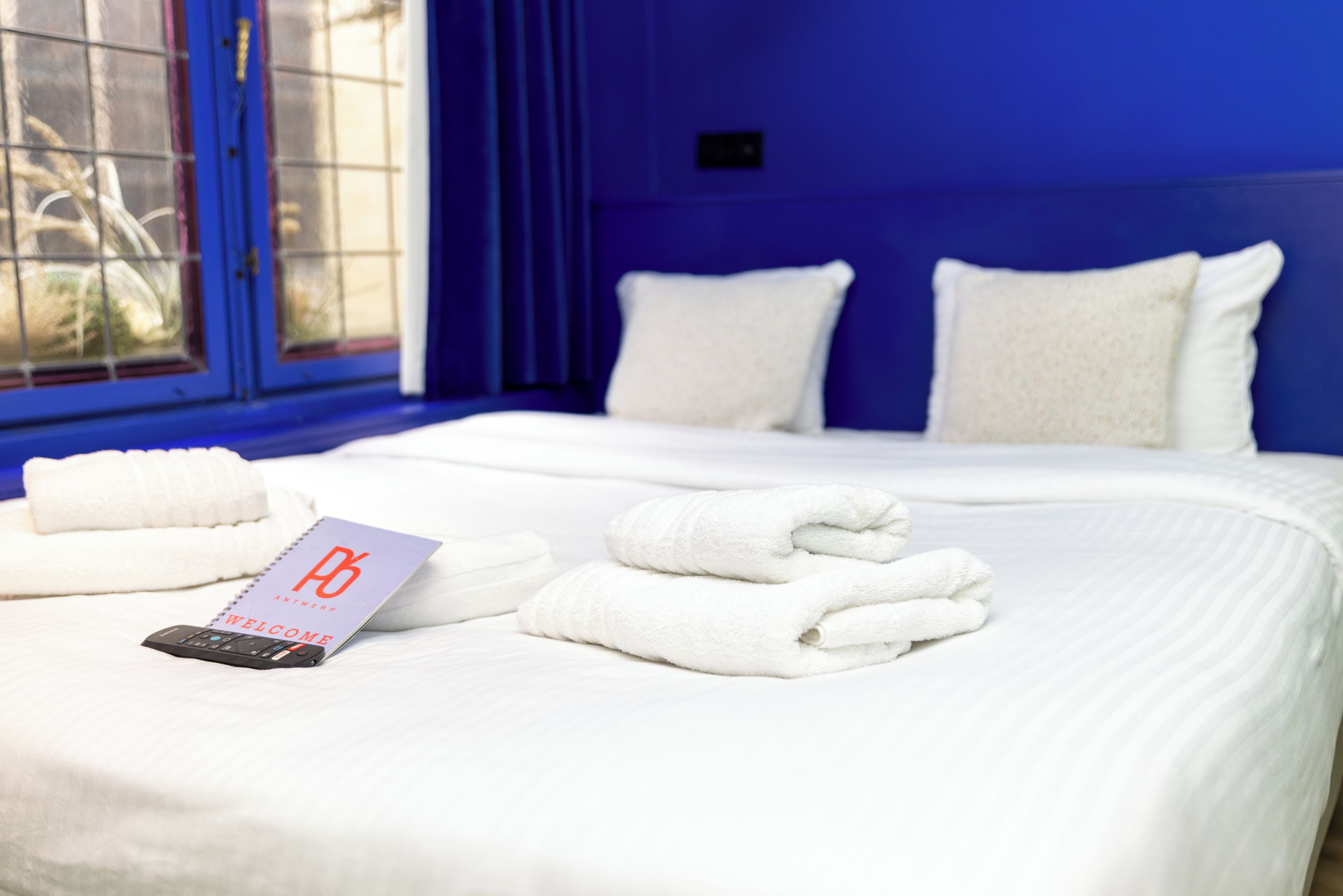 A neatly made bed with white linens and pillows, a welcome card, and a smartphone on the bed in a room with blue walls and a window with a view of plants.