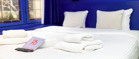 A neatly made bed with white linens and pillows, a welcome card, and a smartphone on the bed in a room with blue walls and a window with a view of plants.