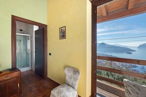 A room with a wooden floor, a chair, a wooden cabinet, a glass shower enclosure, and a large window with a view of mountains and a body of water. - Room 'Stanza 2' with Lake View, Wi-Fi and Air Conditioning (Marone)