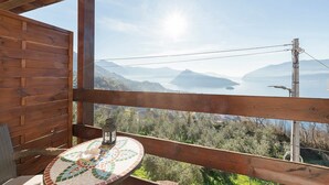 Un balcón de madera con una mesa y un farol, con vistas a un paisaje montañoso y una extensión de agua.