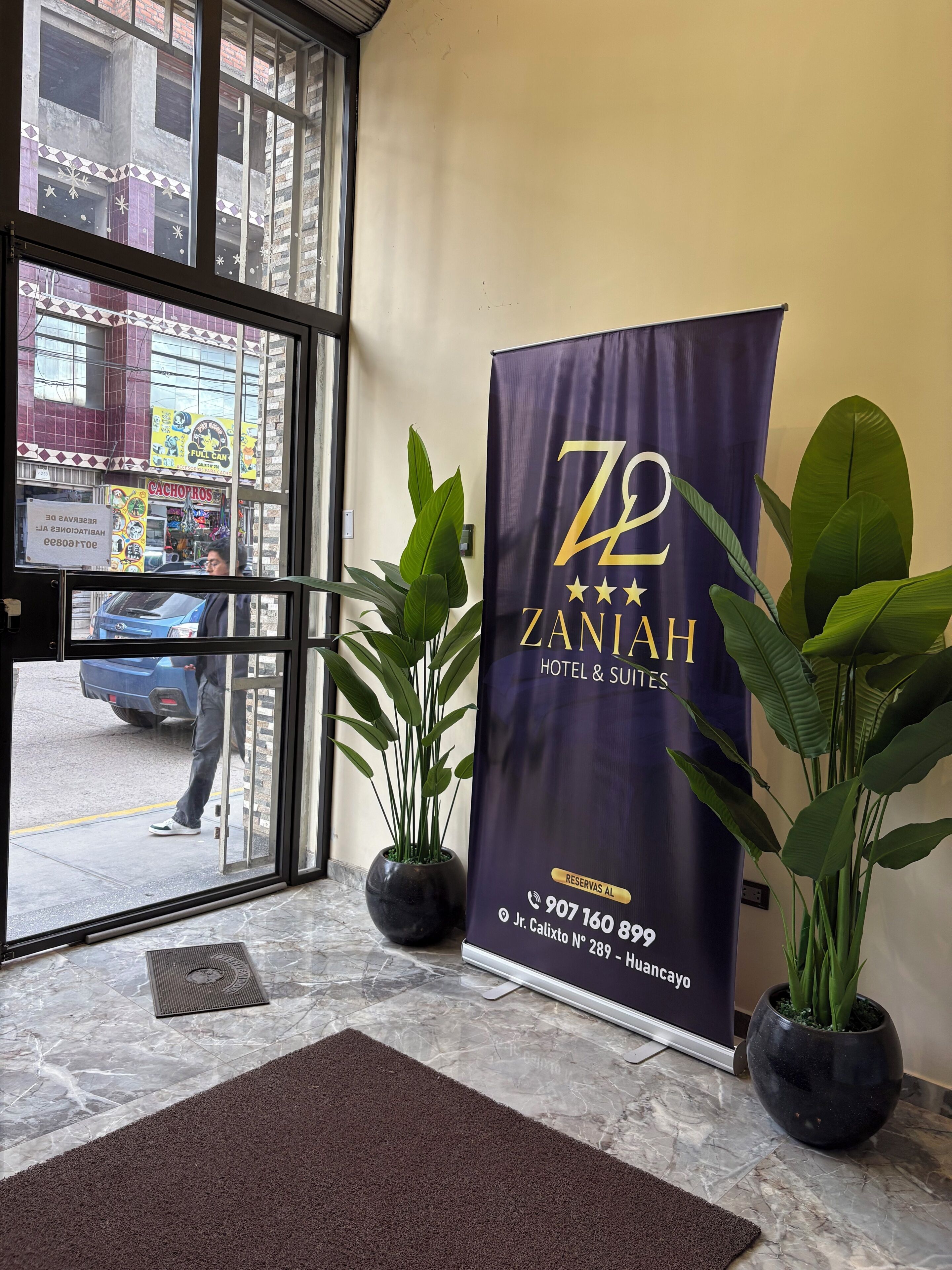 Entrada de un hotel con un cartel de Zaniah Hotel & Suites, una planta en maceta y vista de la calle.