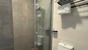 A bathroom with a glass-enclosed shower, a toilet, a black trash bin, and a towel rack with a white towel.